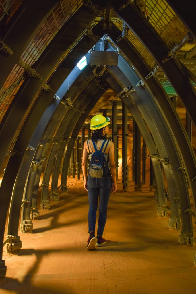 A woman in a hard hat walks through an illuminated industrial tunnel wearing a backpack.