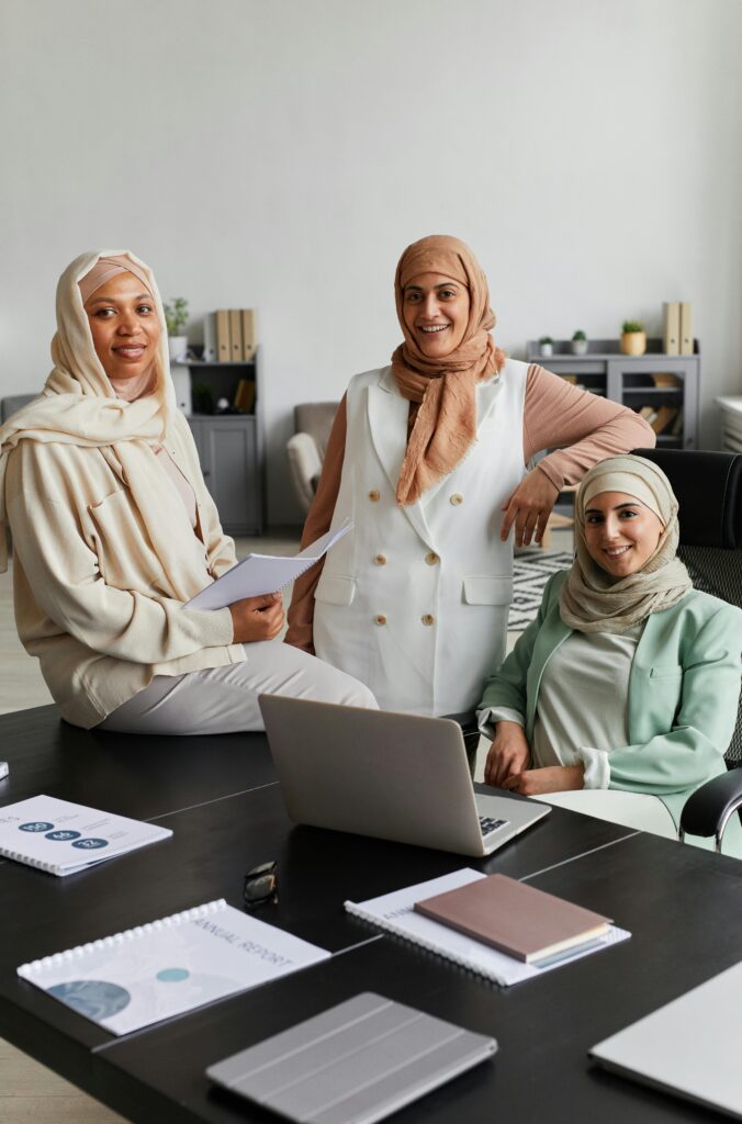 Three Muslim women collaborating in a modern office setting with a laptop.