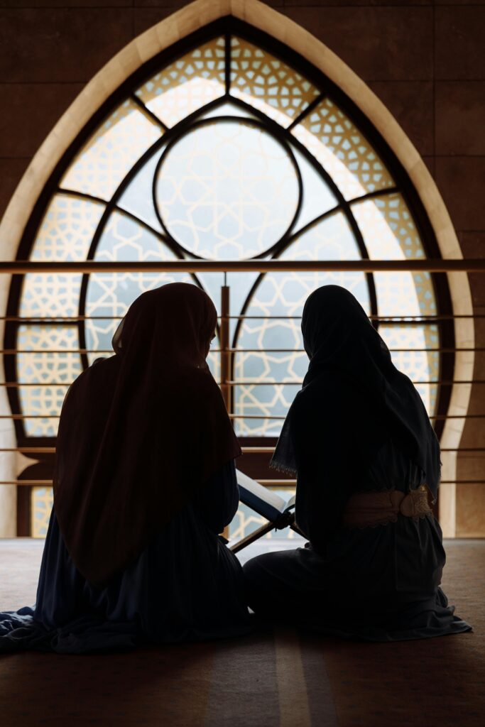 Silhouette of two women in hijab sitting indoors near ornate window in Islamic architecture.